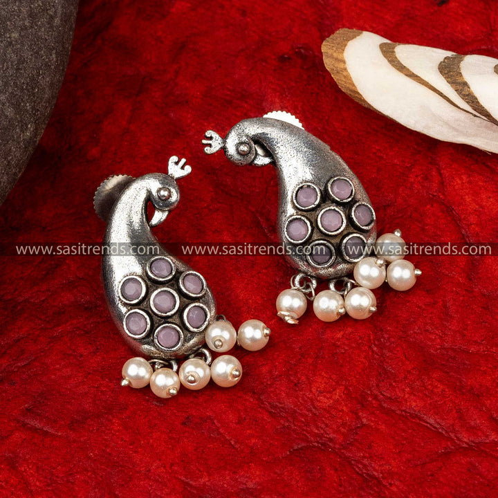 Silver peacock earrings inlaid with purple stones and hanging pearls, displayed on red