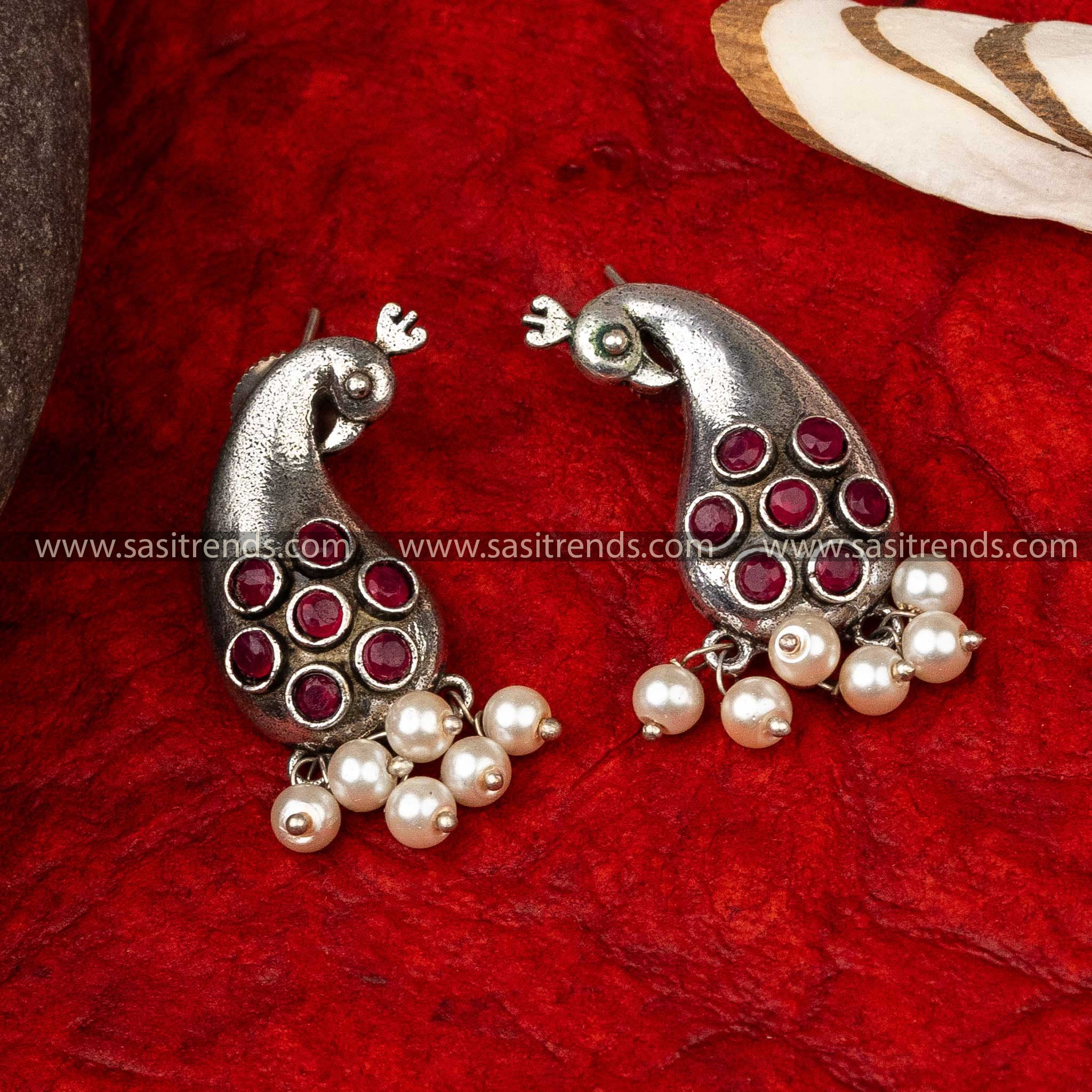 Silver peacock-shaped earrings with circular patterns and pearl drops on a red background